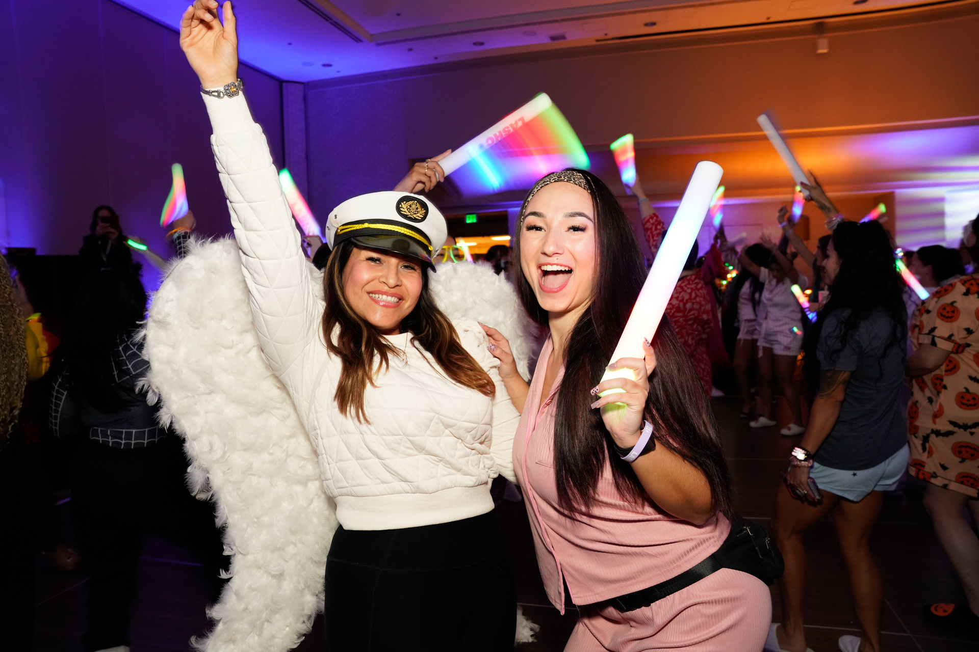 Two people smiling at a party, one with angel wings and a captain's hat, holding glow sticks.