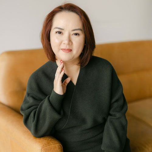 A person with chin-length brown hair wears a dark green sweater while sitting on a tan sofa, resting a hand on their chin.
