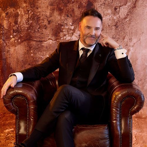 A man in a suit and tie sits relaxed in a tufted brown leather armchair against a textured, reddish-brown background.