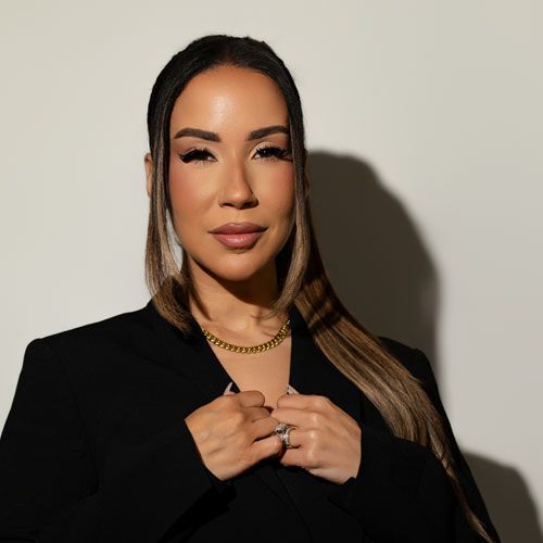 A person with long, two-toned hair wears a black blazer and gold chain, holding their hands together against a gray wall.
