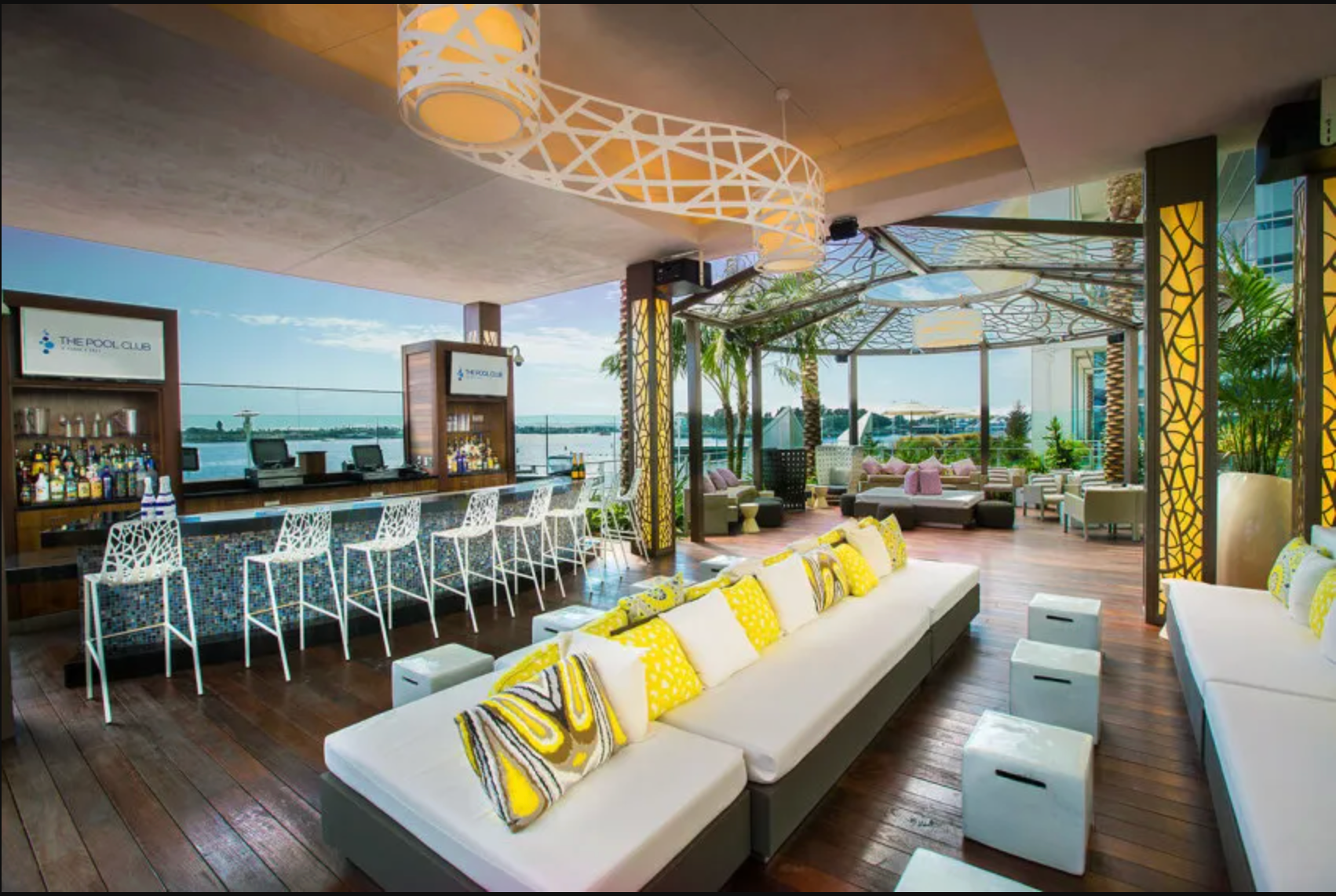 An outdoor lounge with a bar, white bar stools, and long sofas with yellow and white pillows overlooking a body of water.