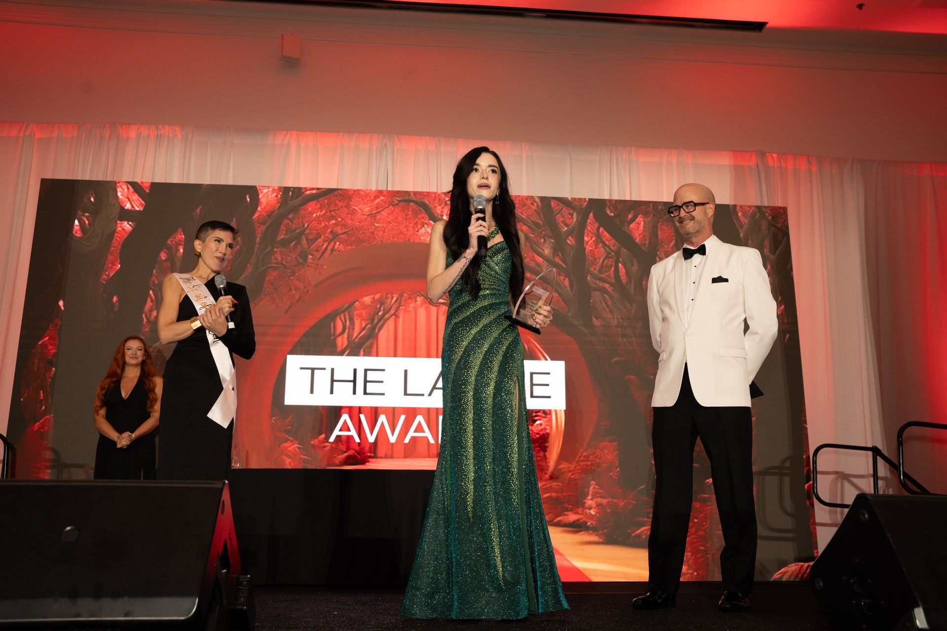 Award ceremony stage with woman in green gown speaking beside two presenters and a large award backdrop