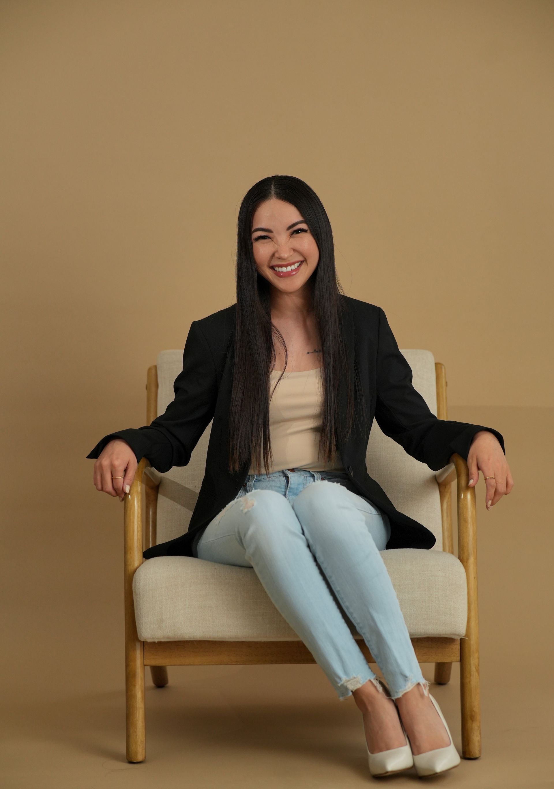 Woman smiling in a black blazer, seated in a beige armchair against a plain tan wall.