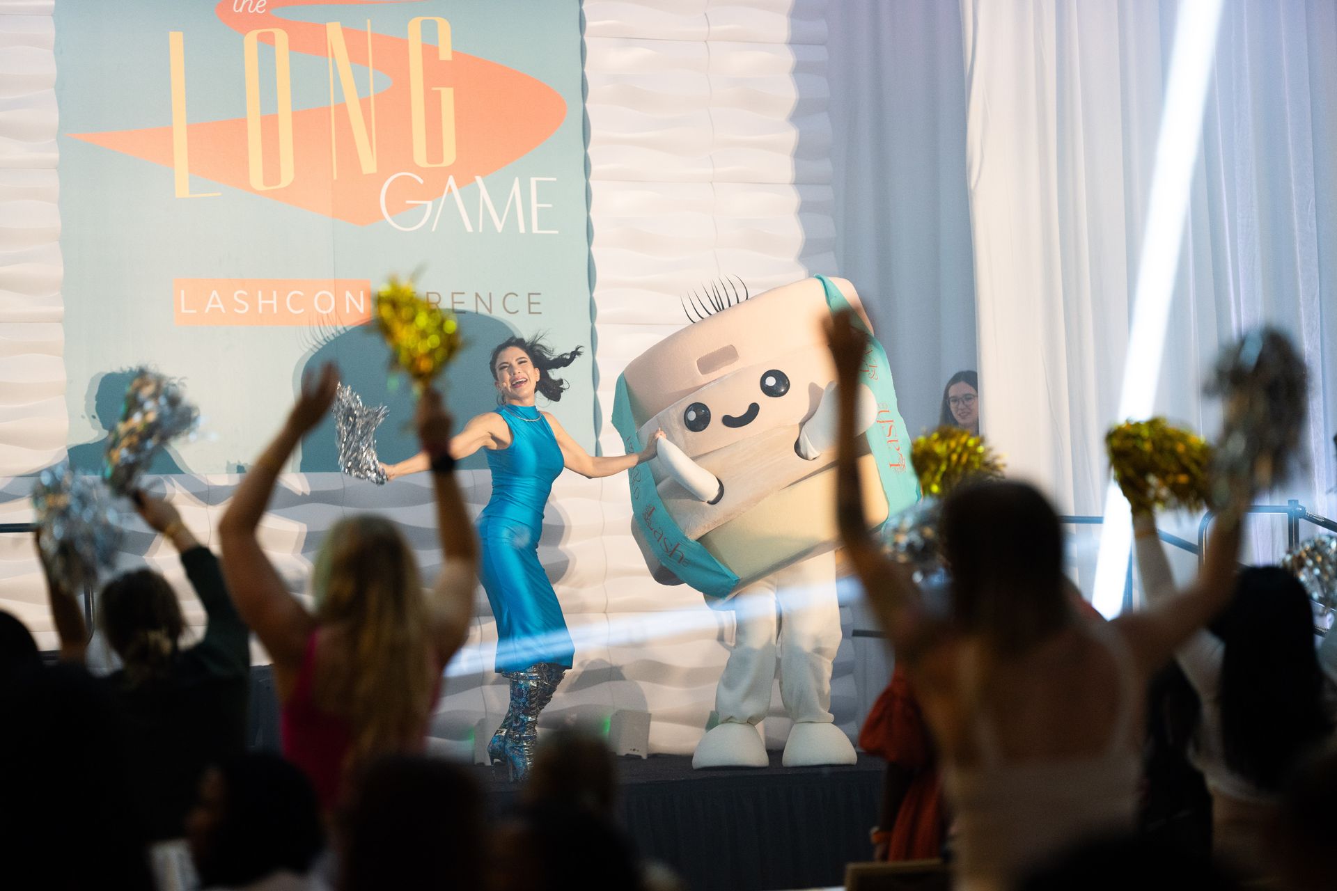 A woman in a blue dress is dancing with a mascot in front of a crowd of people.
