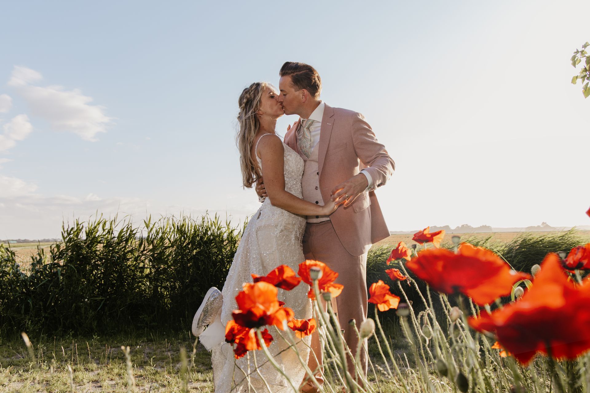 Bruid en bruidegom kussen elkaar in een zonovergoten veld met felrode bloemen op de voorgrond.