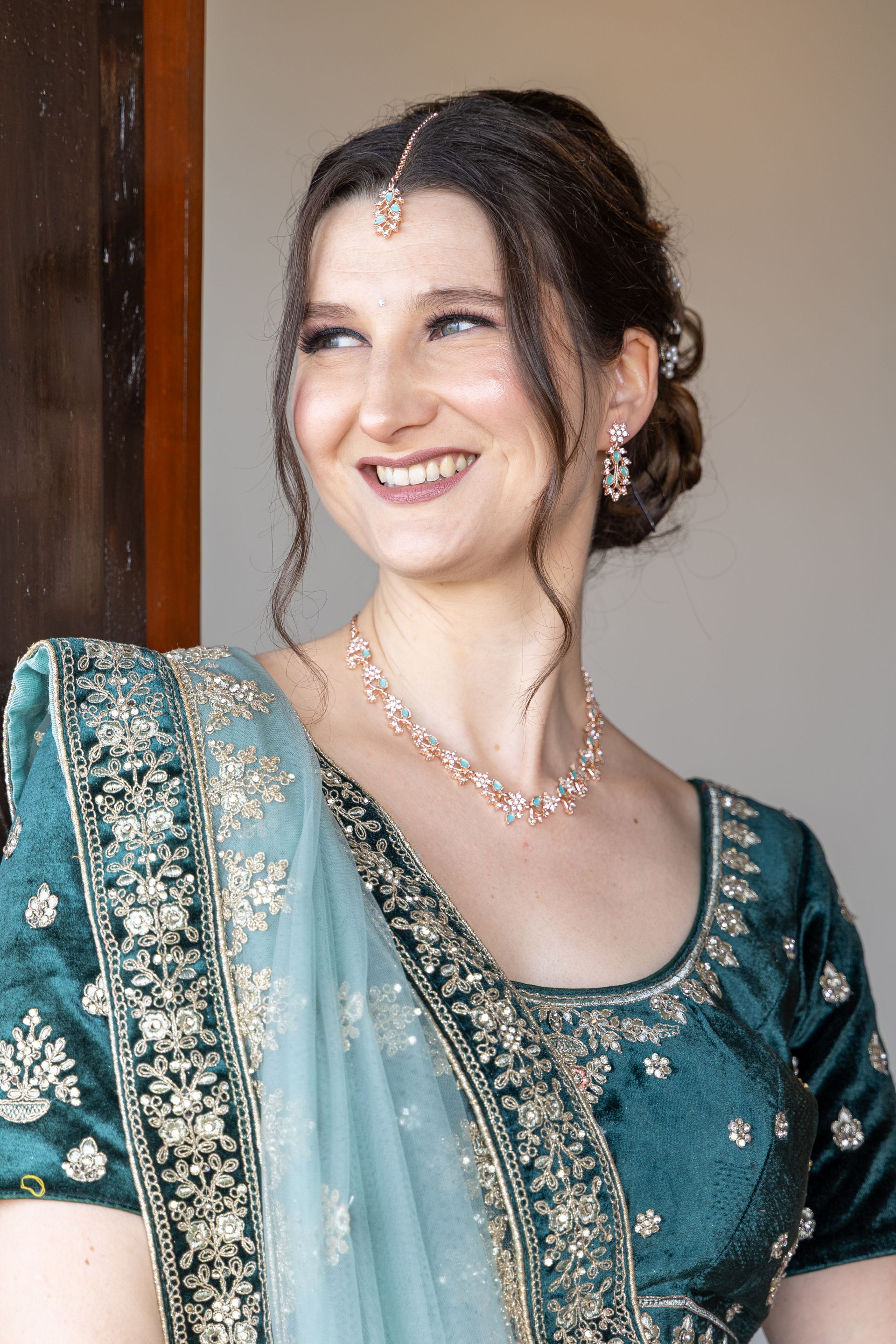 Een lachende vrouw in een turquoise geborduurde jurk en sieraden staat binnen bij een deuropening.