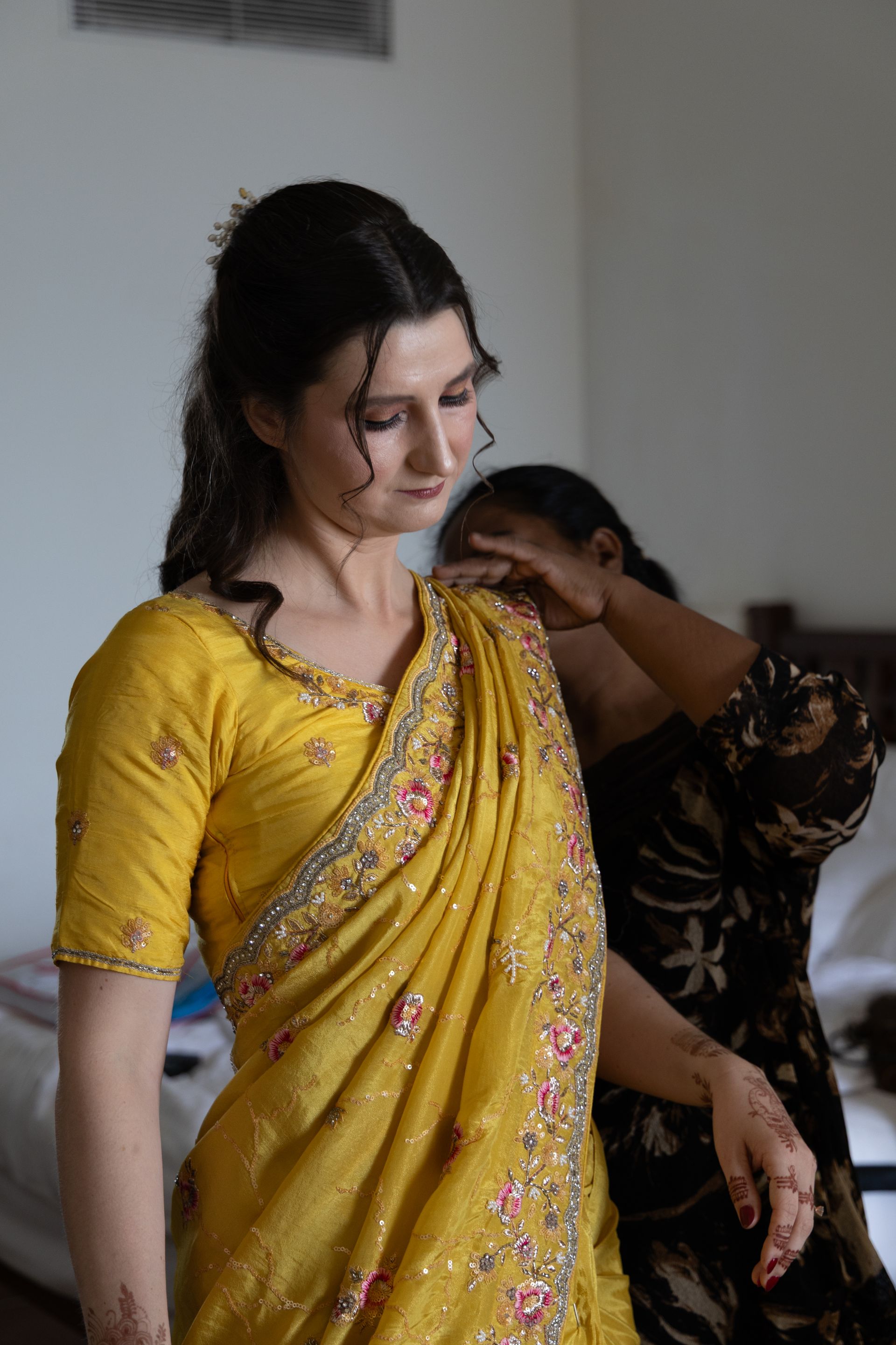 Vrouw in een gele sari wordt binnenshuis door iemand anders geholpen bij het in orde maken van haar sari.