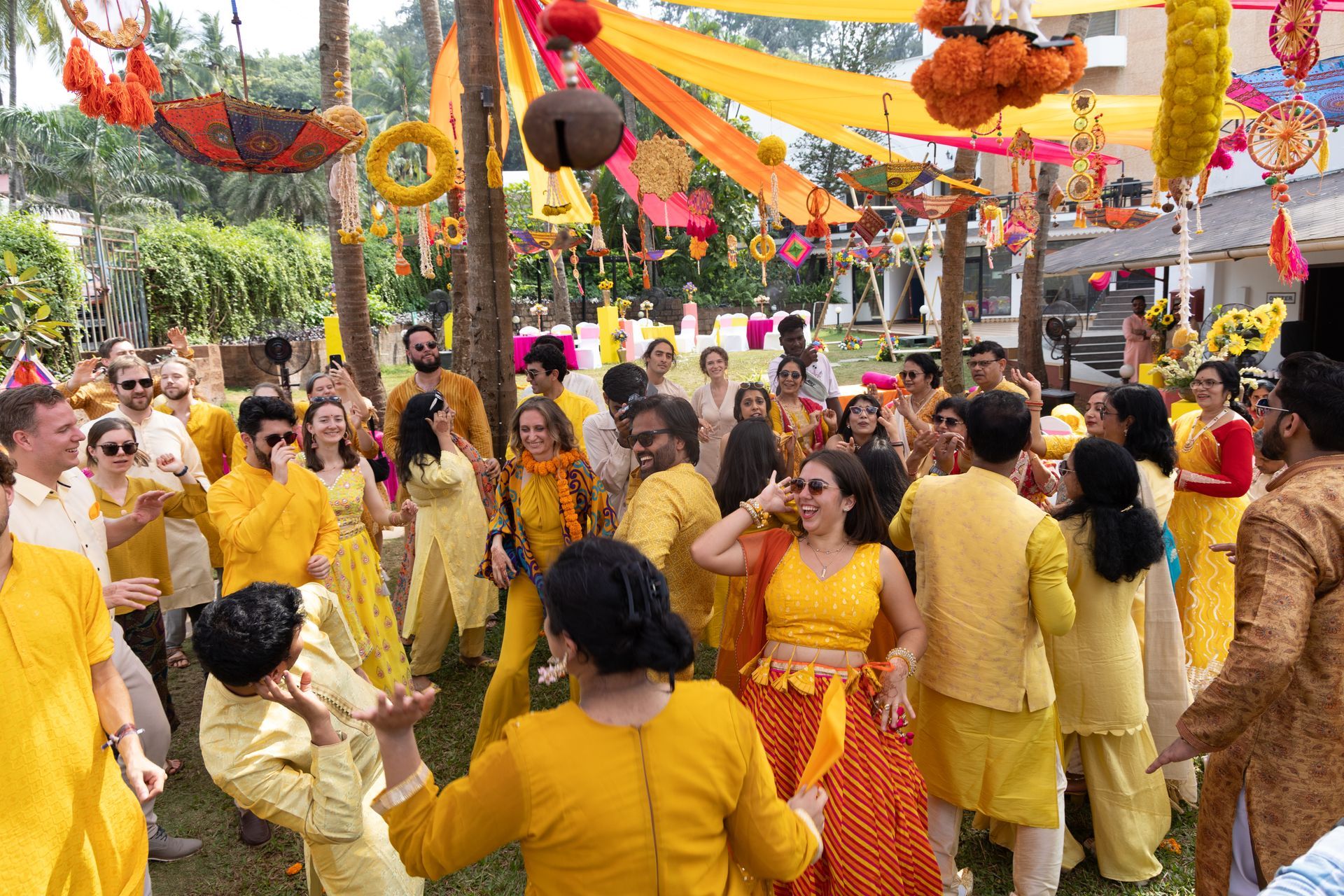Een menigte in gele en oranje kleding danst op een feestelijk buitenfeest met kleurrijke versieringen boven hun hoofden.