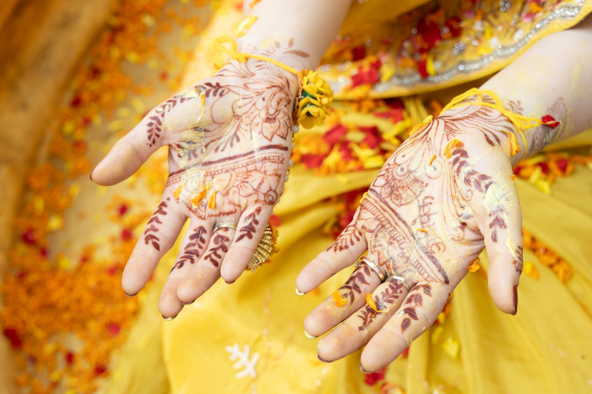 Twee handen met ingewikkelde henna-ontwerpen op felgele stof en goudbloemblaadjes.