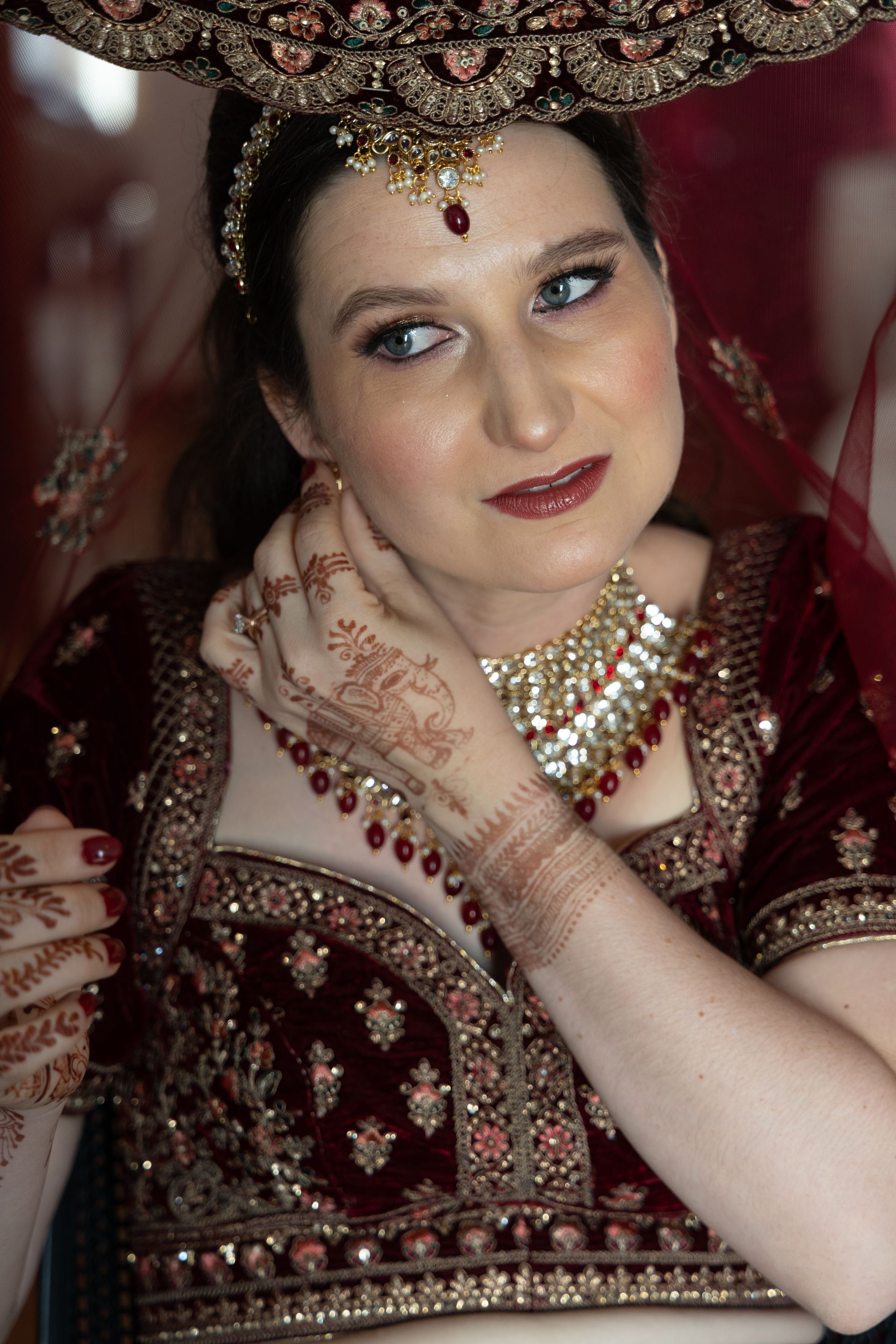 Vrouw in een rijk versierde rode bruidsjurk verstelt haar halsketting, draagt een met juwelen bezette hoofdtooi en heeft met henna versierde handen.
