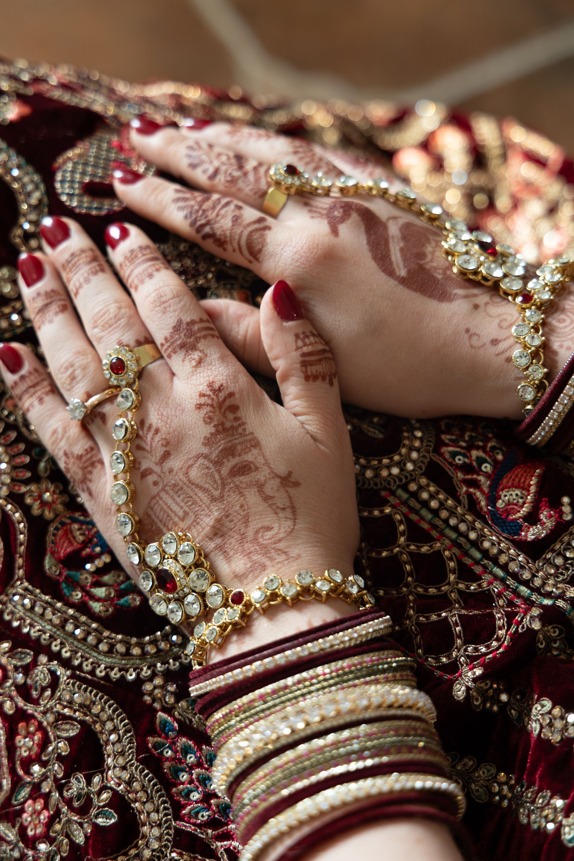Versierde handen met henna, rode nagels en gouden armbanden rusten op een geborduurde bordeauxrode stof.