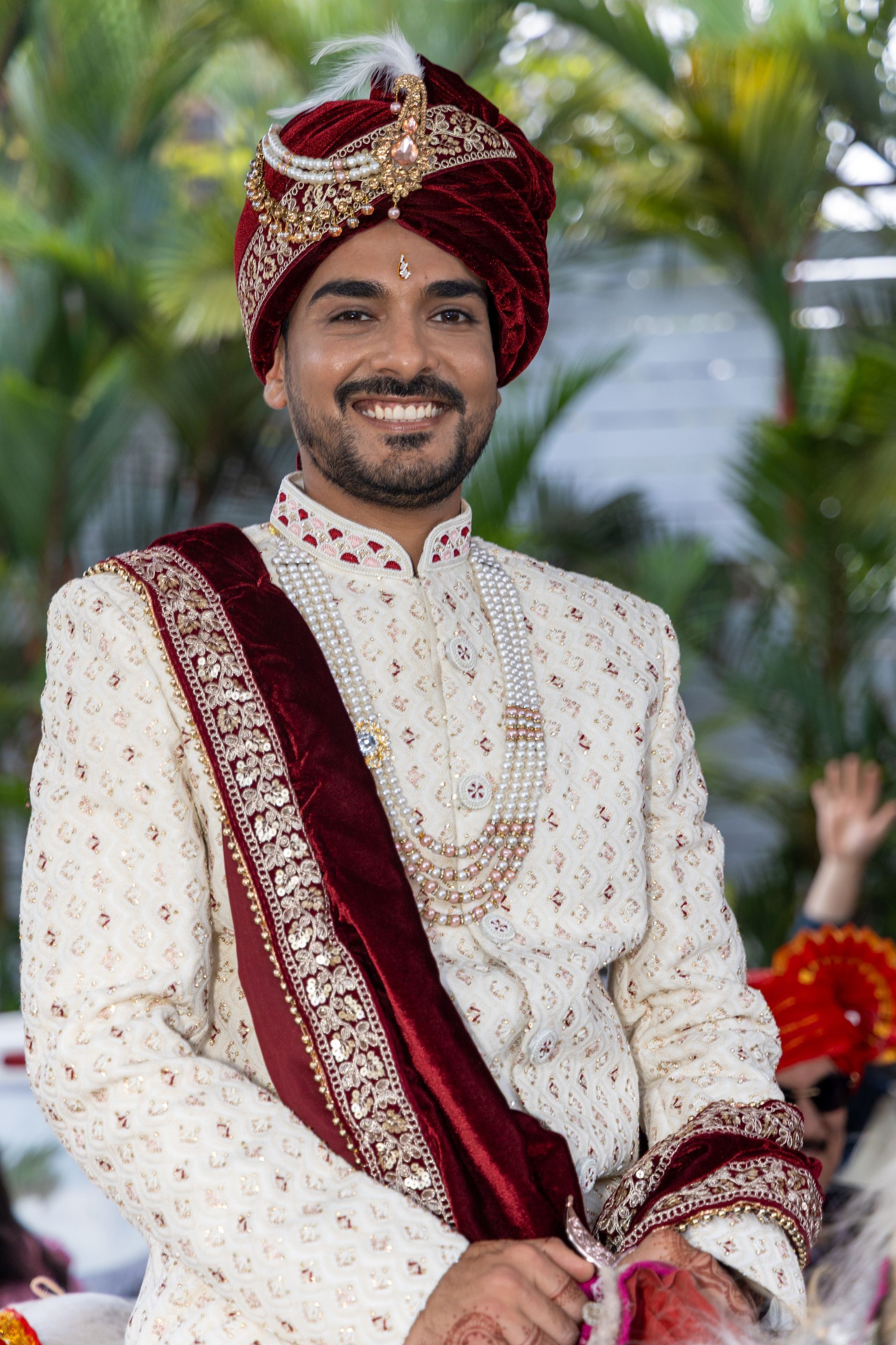 De bruidegom, stralend van plezier, gekleed in een witte sherwani en een bordeauxrode tulband, zit binnen met een bloemenachtergrond.