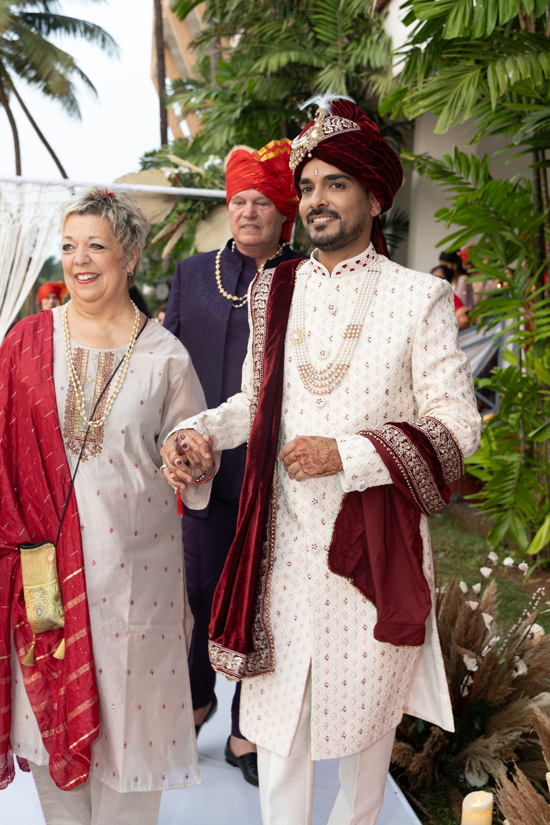 Drie mensen in rijkversierde traditionele kleding wandelen buiten tussen weelderig groen.