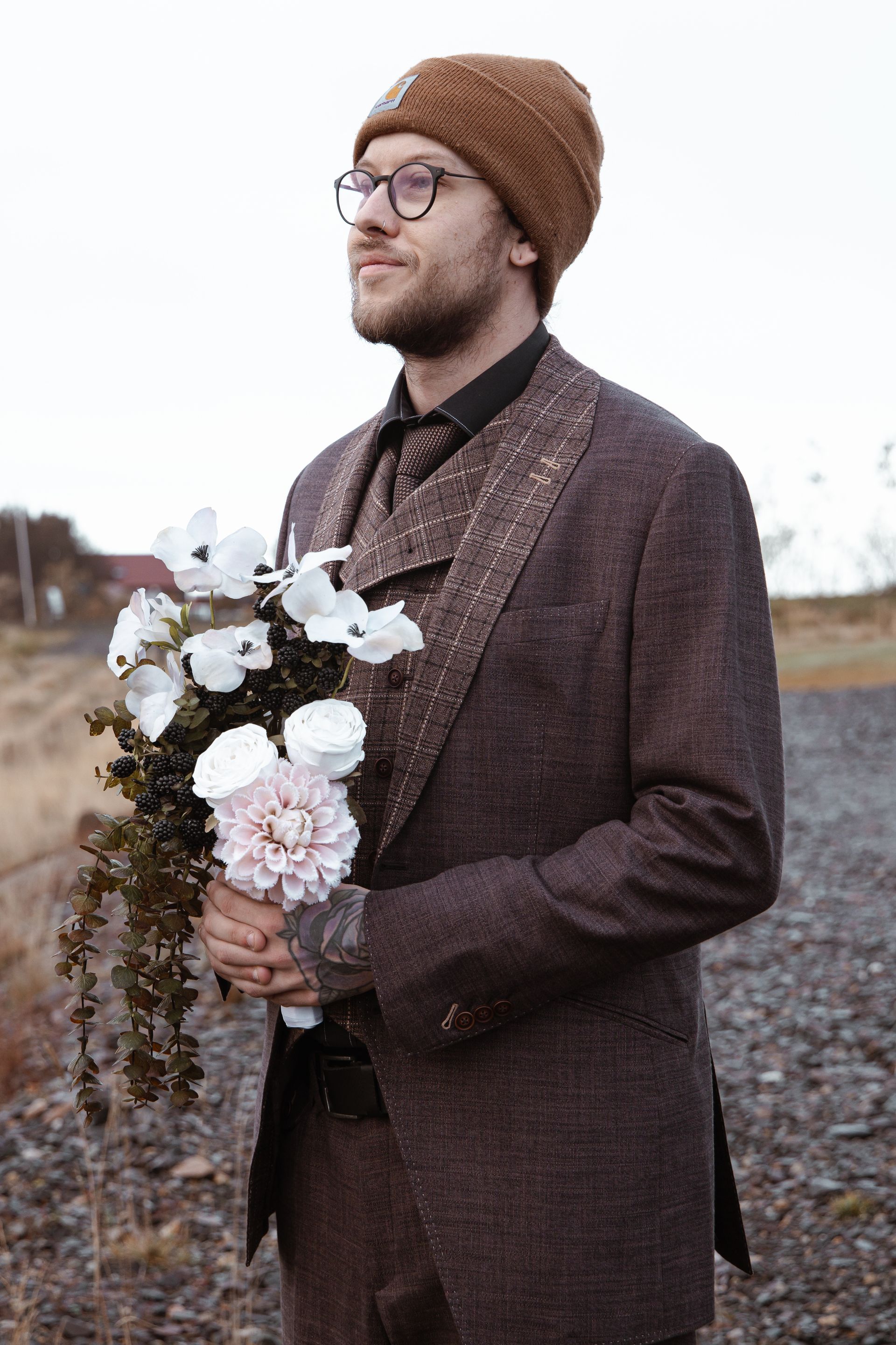 Man in bruine jas en gebreide muts met een boeket witte bloemen buiten op een bewolkte dag.