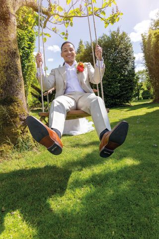 Een persoon zit op een schommel in een zonnige tuin, gekleed in een wit pak met oranje bloemen en zwarte schoenen.