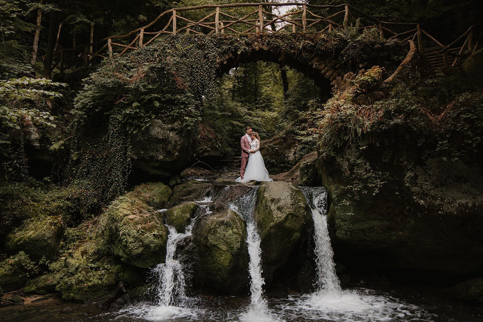 Een persoon in een witte jurk staat op rotsen bij een kleine waterval in een bos.