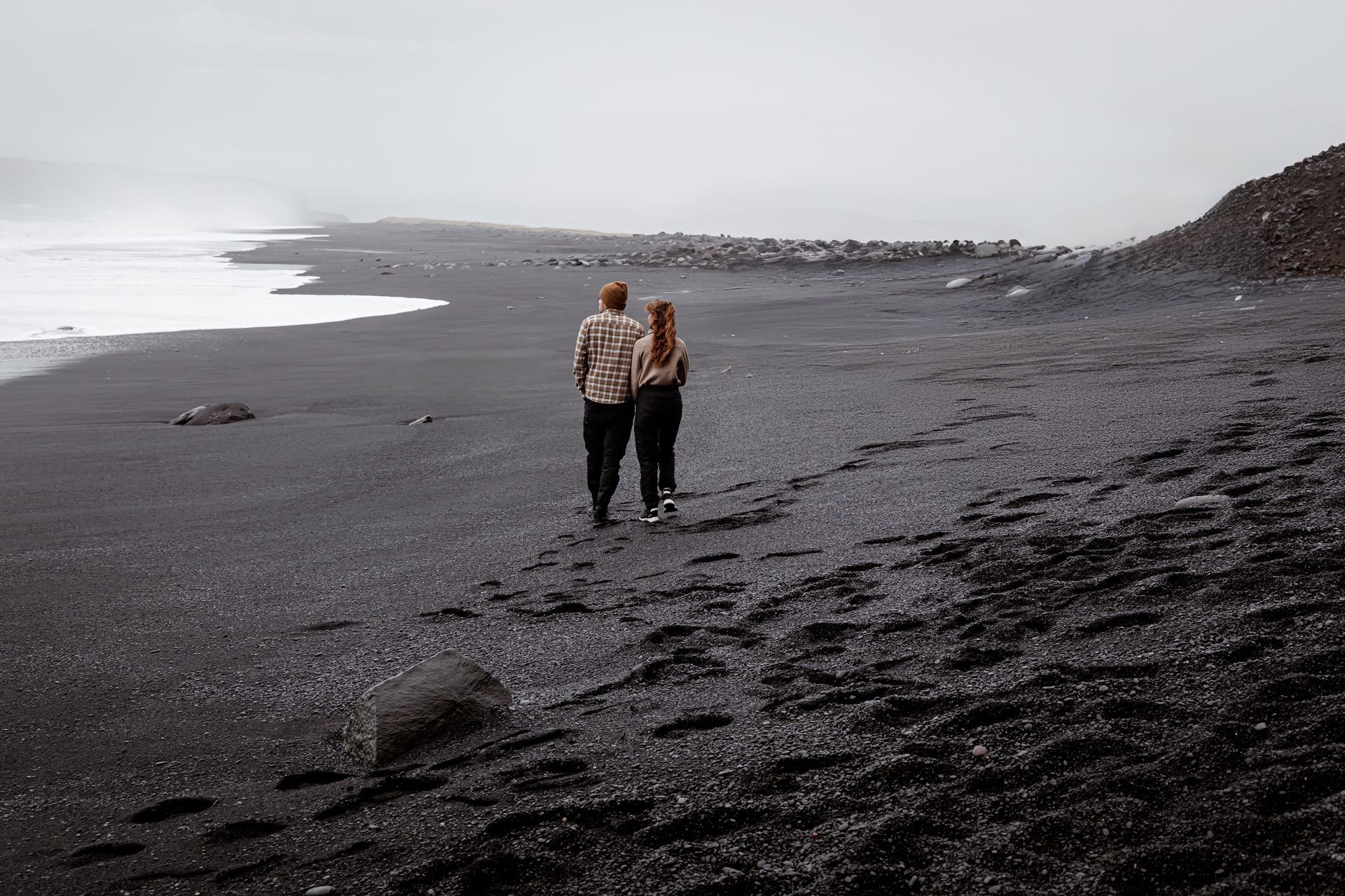 Twee mensen lopen over een donker, rotsachtig strand langs kalm water onder een bewolkte hemel.
