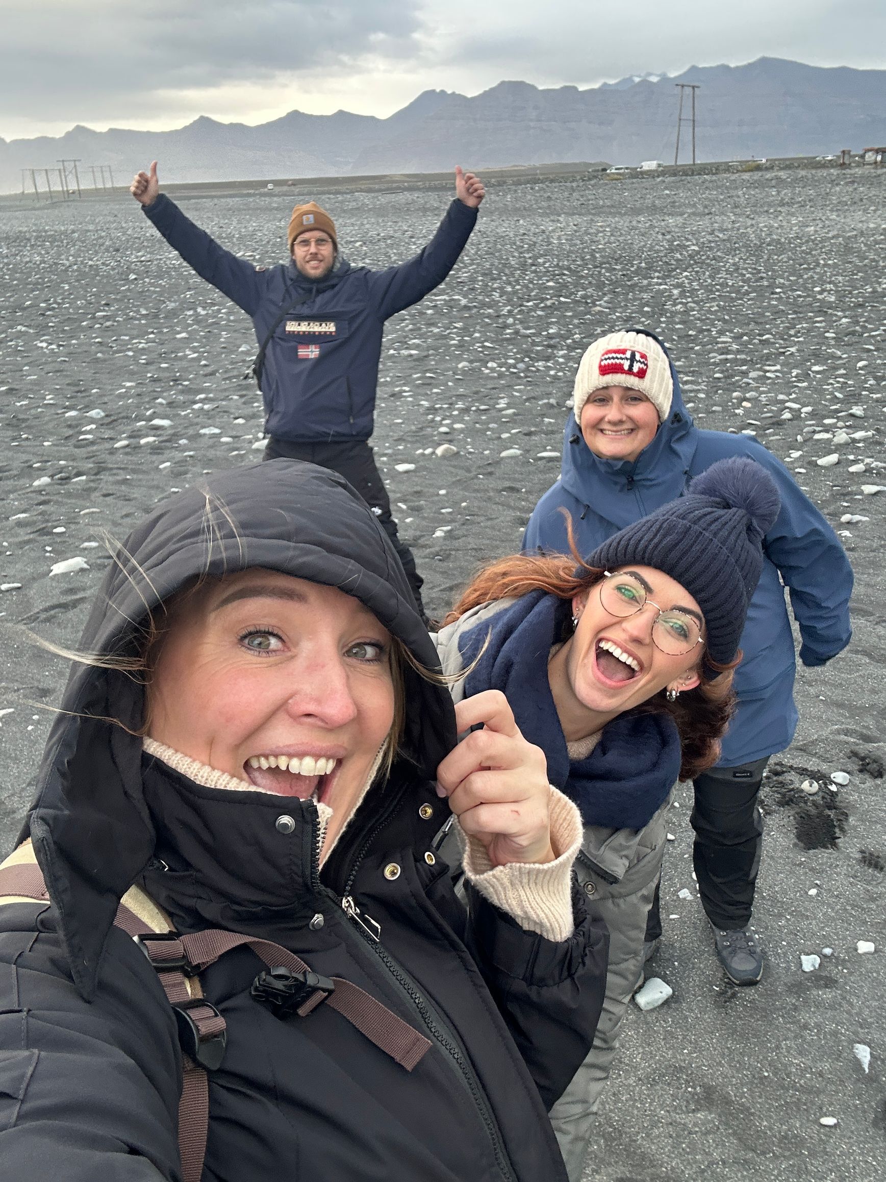 Vier mensen lachend op een winderig, rotsachtig strand, waarvan één een selfie maakt met bergen op de achtergrond.