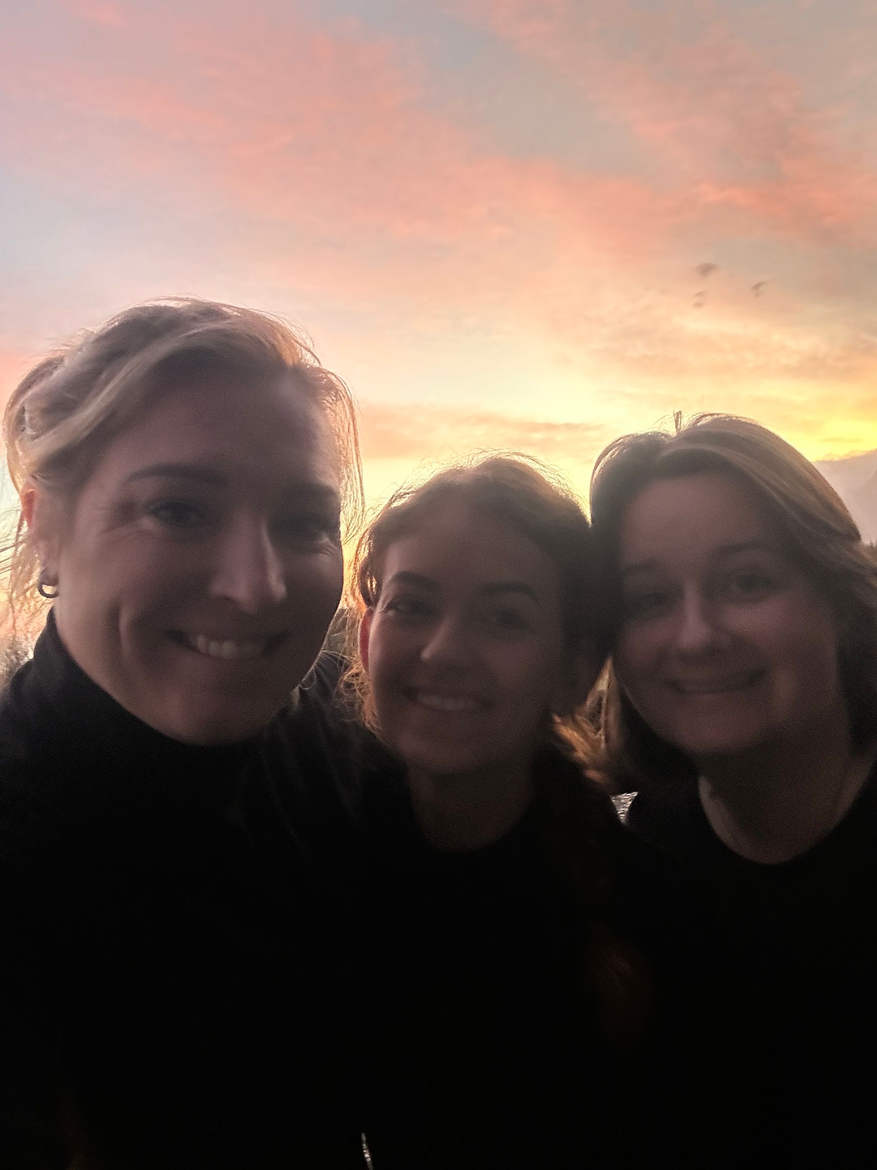 Drie mensen glimlachen buiten bij zonsondergang onder een roze-oranje hemel.