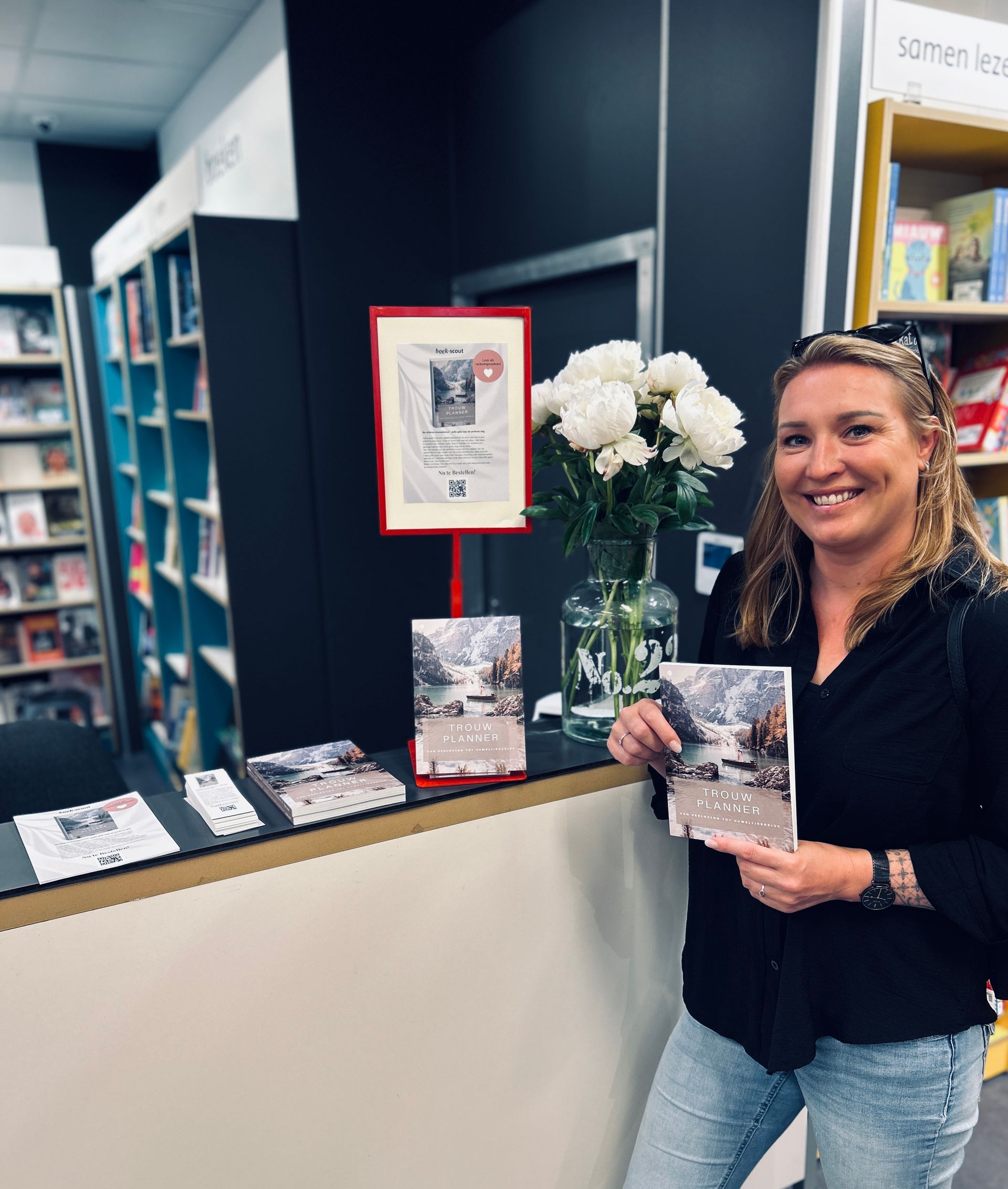 Een vrouw lacht naast een boekendisplay in een boekwinkel, met een boek in haar handen, omringd door bloemen en ingelijste borden.