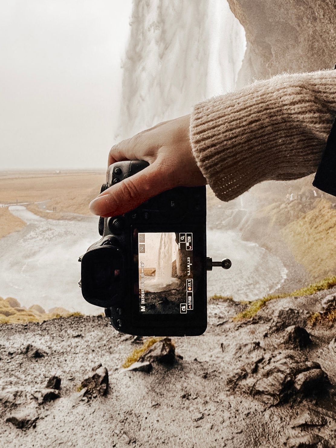 Een hand houdt een camera vast op een natte, rotsachtige kust naast een waterval en een mistige klif.