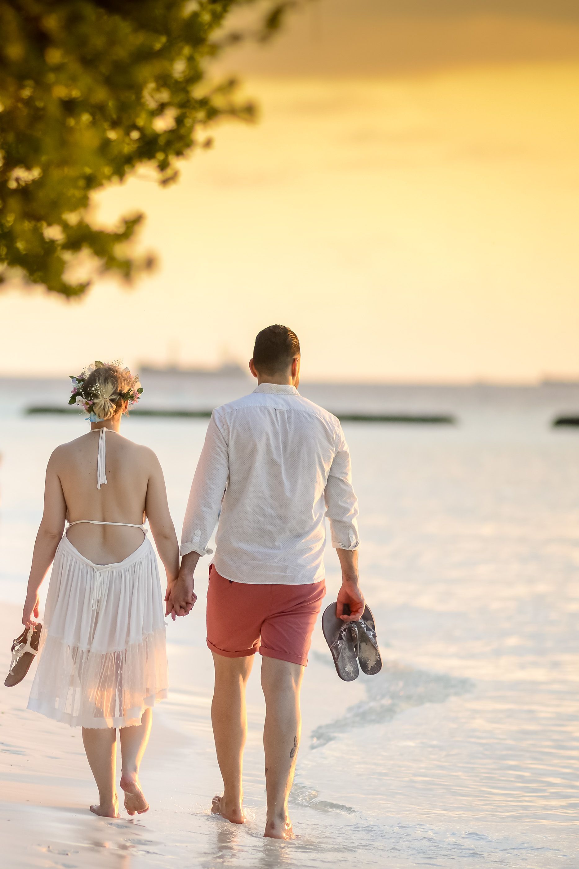 Een stel loopt op blote voeten over het strand bij zonsondergang, hun schoenen vasthoudend, met kalm water en een gouden hemel.