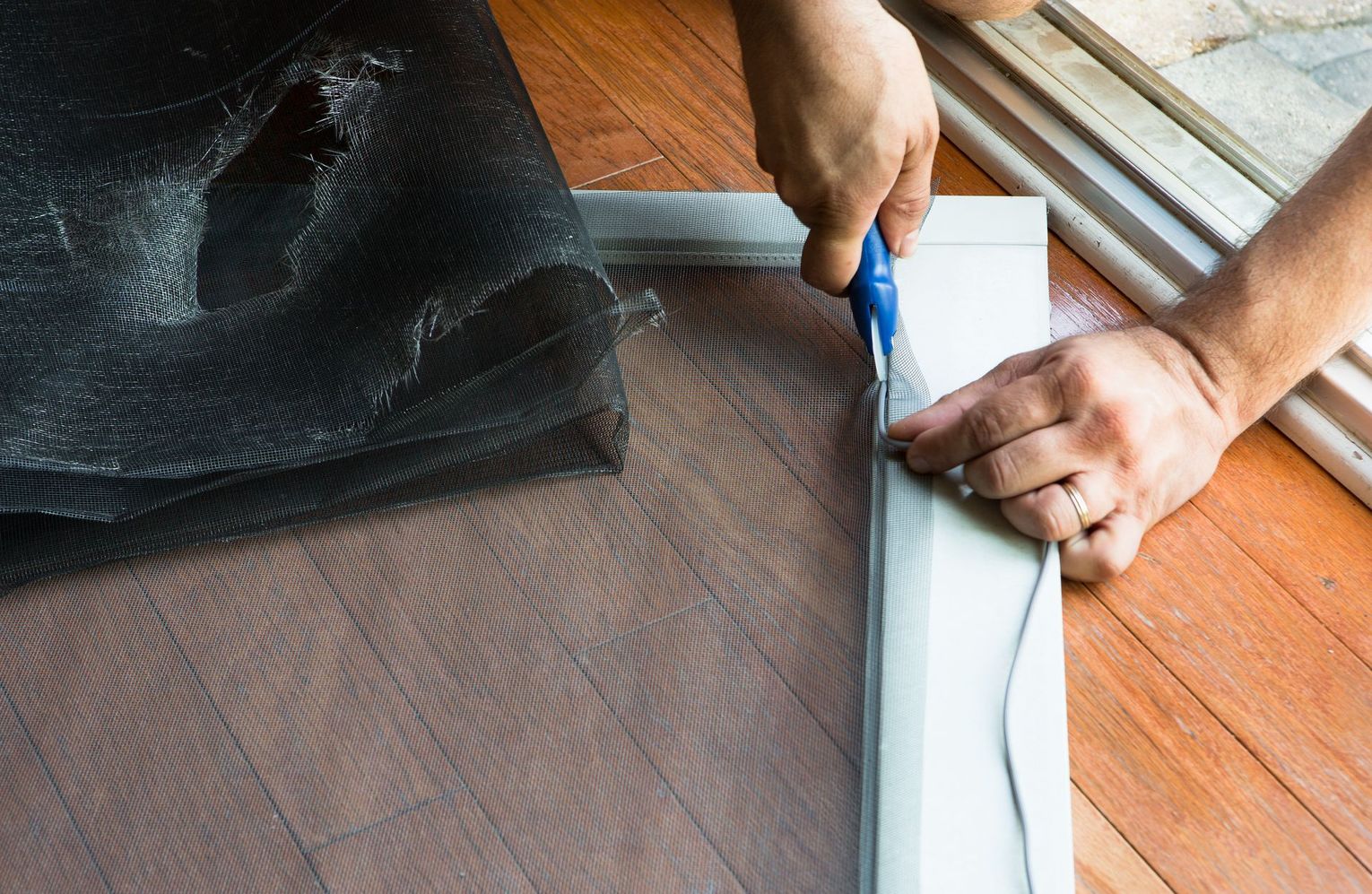 Hands cutting black mesh fabric with blue scissors on a wooden floor