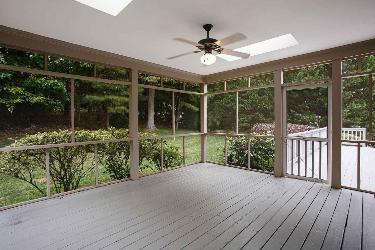 Screened porch with gray floor, ceiling fan, skylights, and wooded view outside.