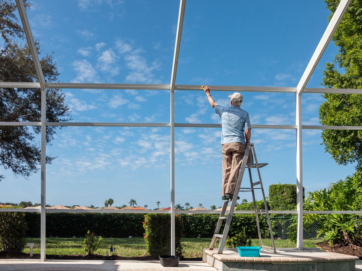 Person on a ladder painting the top of a white screened patio frame under a blue sky
