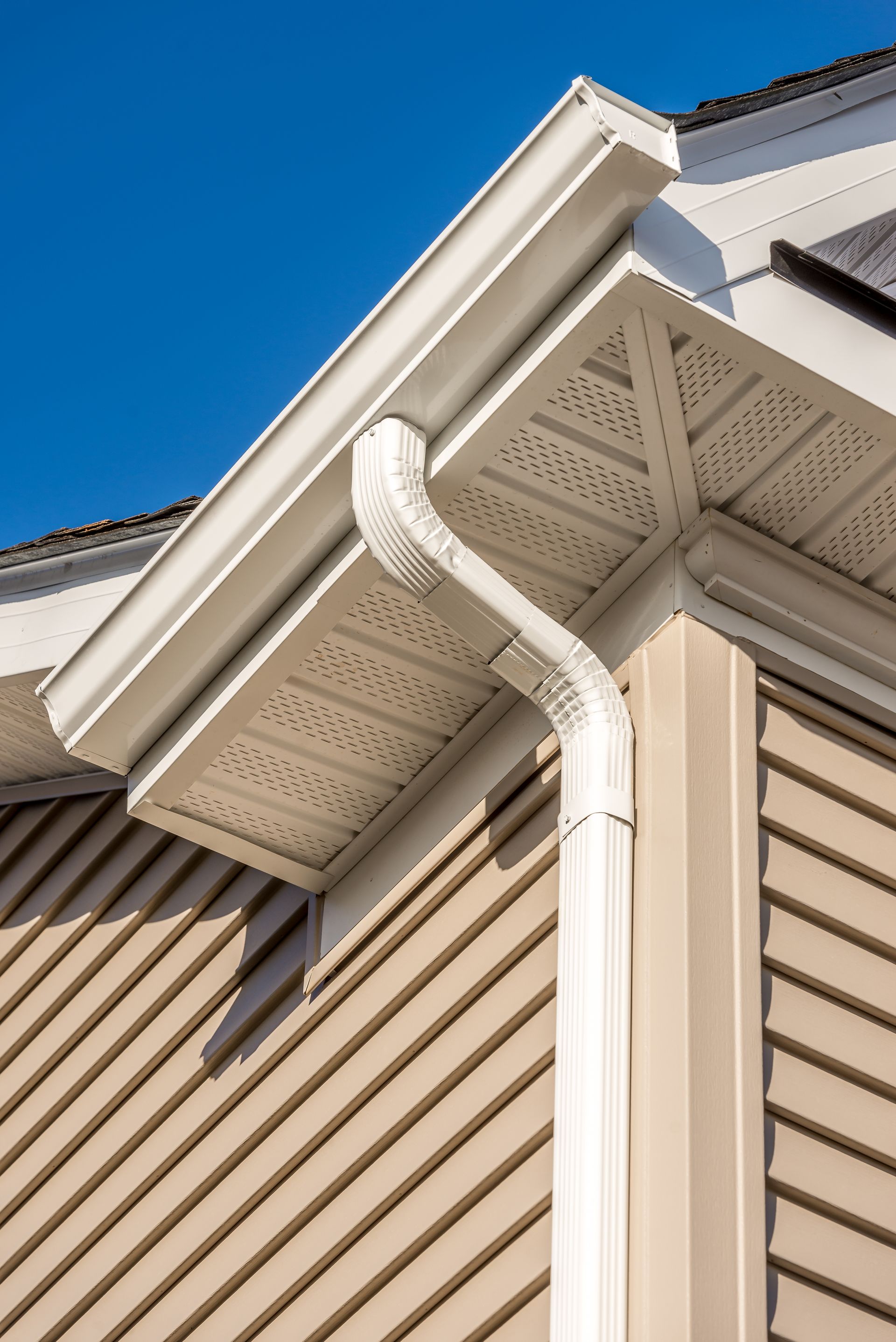 White rain gutter and downspout on beige house siding under a bright blue sky