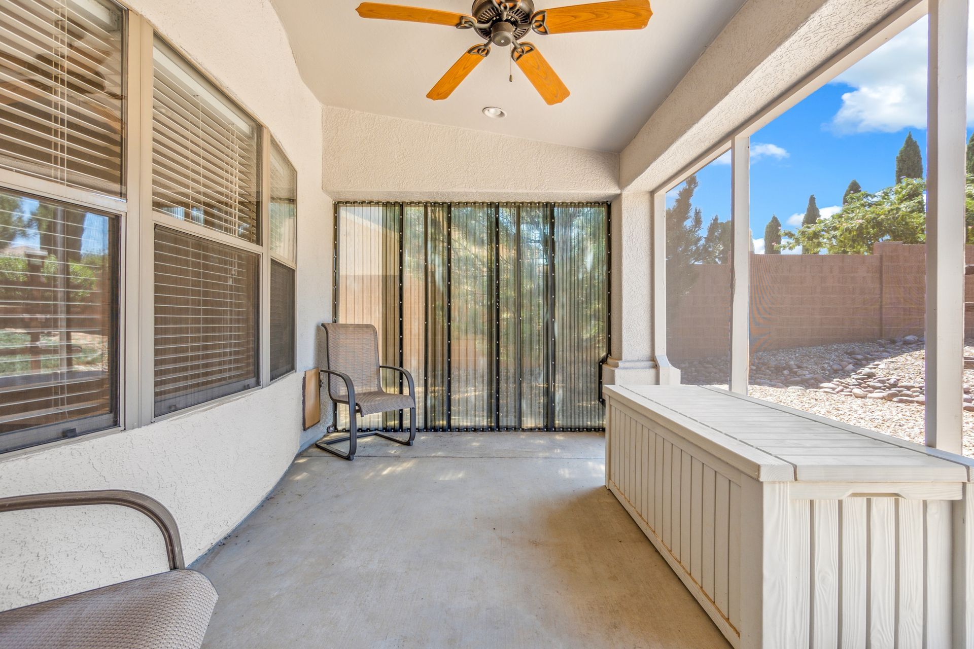 Covered patio with ceiling fan, windows, sliding glass door, and desert landscaping beyond the fence