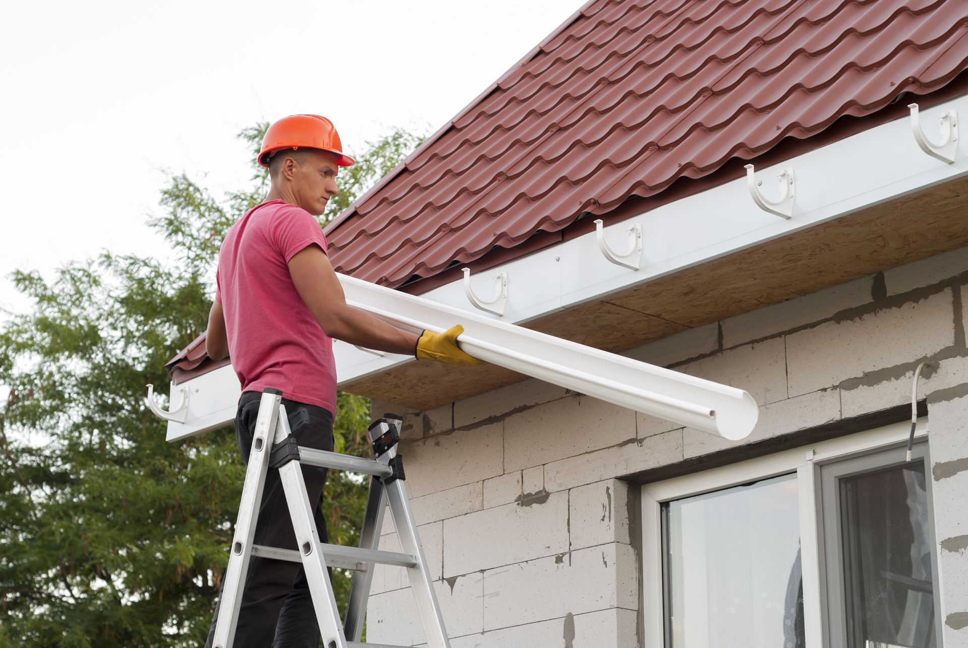 Worker on a ladder installing white guttering under a red-tiled roof