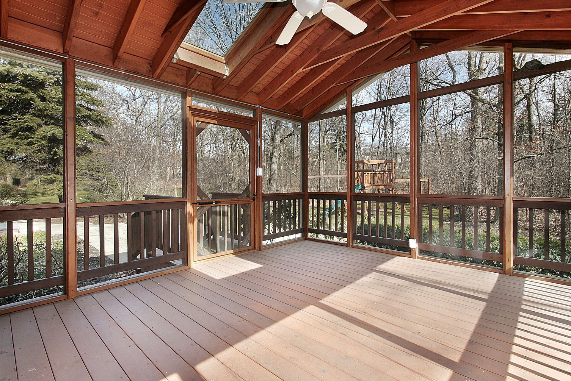 Sunlit screened porch with wooden floor, railing, and ceiling fan overlooking trees