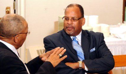 Two Black men in suits engaged in conversation. One gestures, other listens attentively, indoors.
