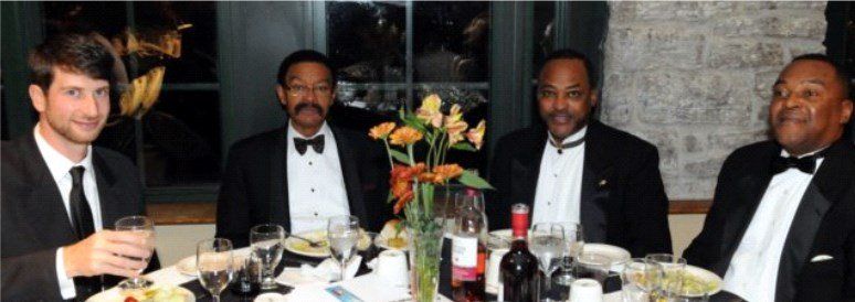 Four men in tuxedos at a formal dinner table, smiling, with flowers and wine.