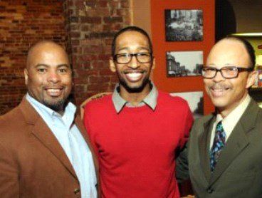 Three smiling Black men pose for a photo indoors. Two wear suits, the center one a red sweater.