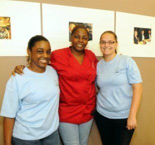 Three women smiling: one in red top, arms around two women in blue shirts, standing in front of photos.