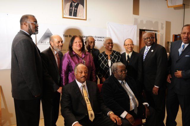 Group of formally dressed people, some seated, pose together indoors; banner in background.