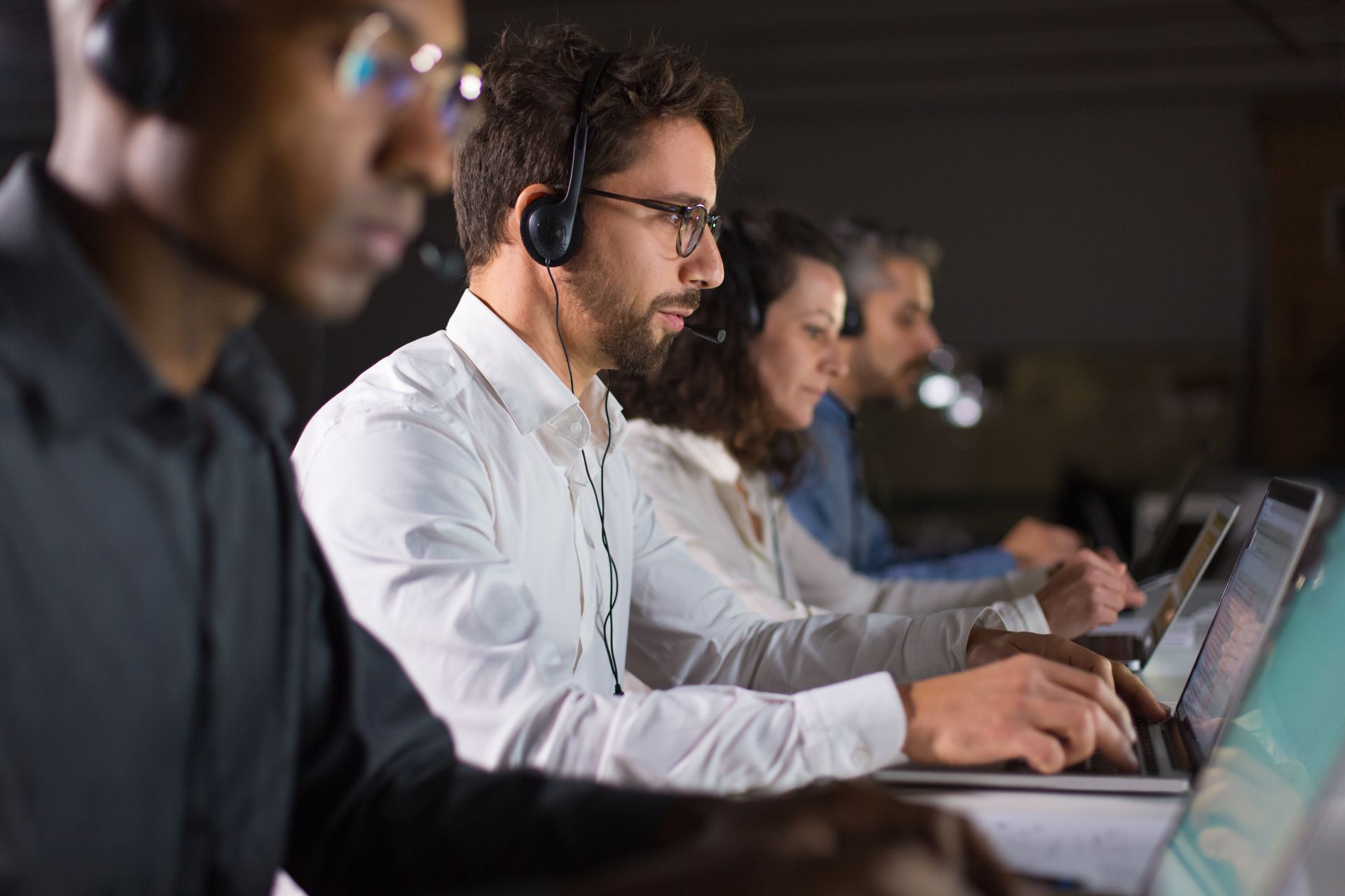 Pessoas usando fones de ouvido trabalhando em laptops em um escritório com pouca luz.