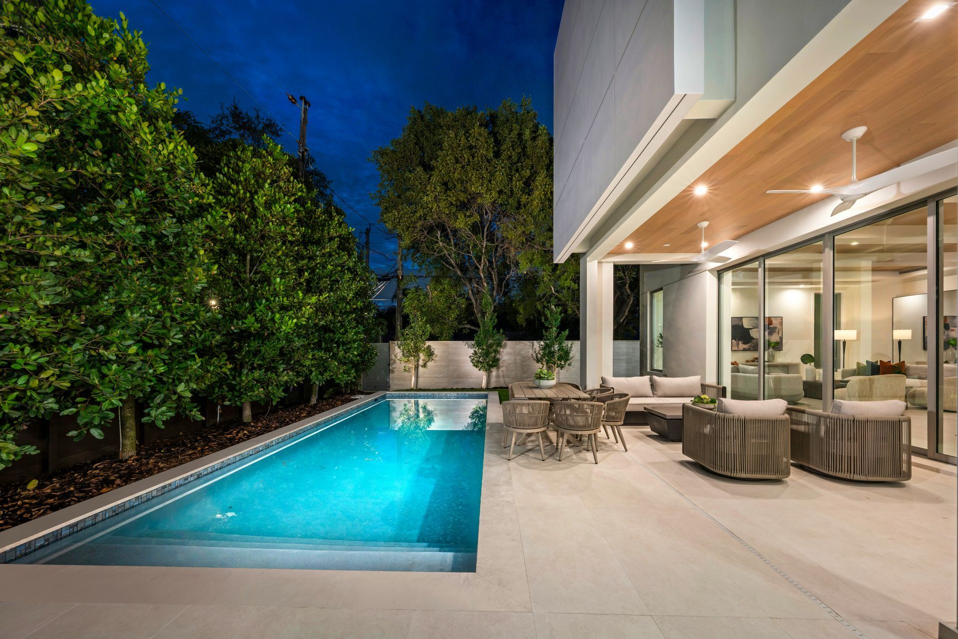 Modern backyard patio with a lit swimming pool, lounge chairs, and a covered outdoor dining area at dusk