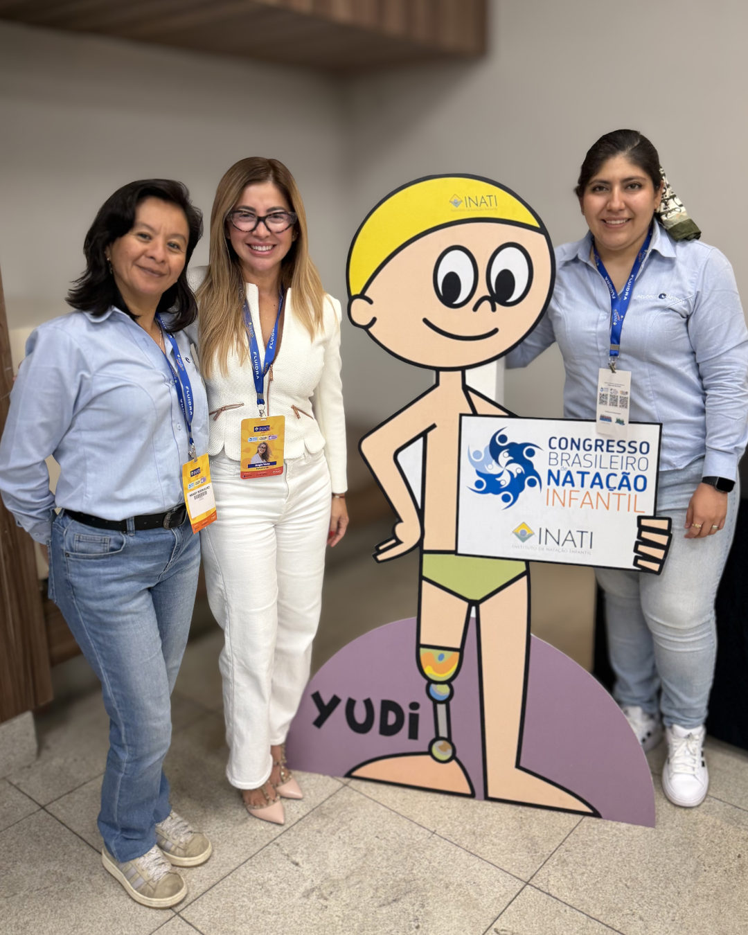 Tres mujeres de pie junto a una figura de cartón de un dibujo animado de Yudi que sostiene un cartel de la conferencia en el interior de un edificio.