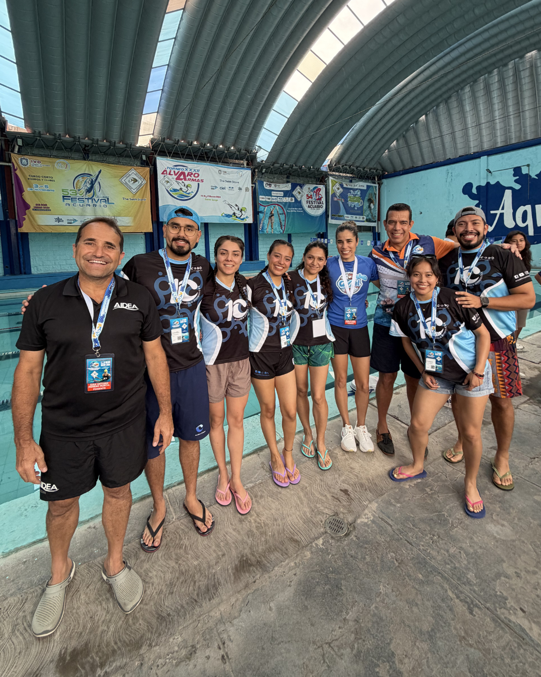 Grupo de personas con camisetas negras del evento posando en el interior, bajo un techo curvo, en un recinto deportivo.