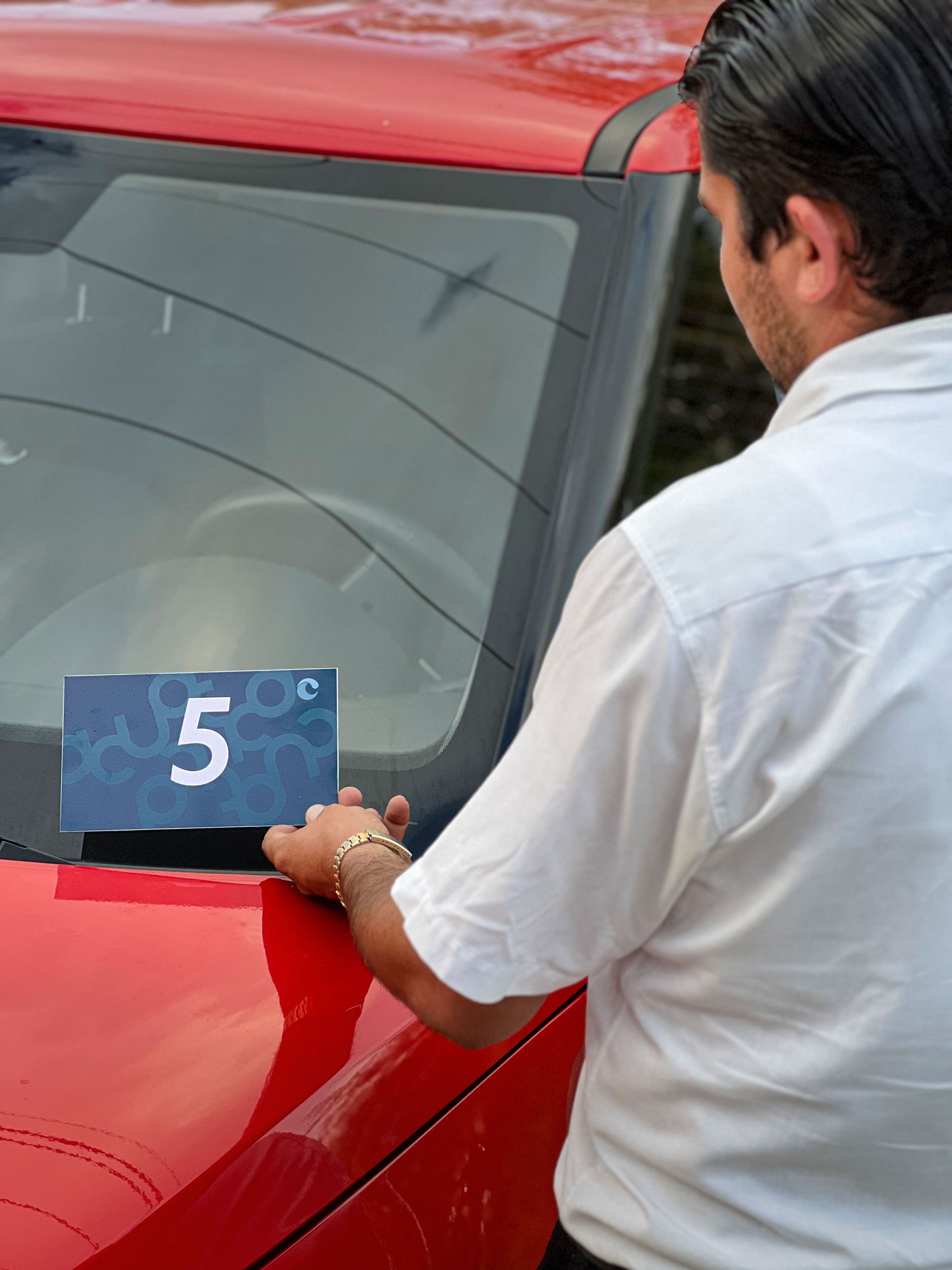 Persona colocando una tarjeta azul con el número 5 en el parabrisas de un coche rojo.