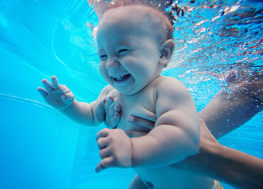 Un bebé sonriente bajo el agua en una piscina, sostenido por la mano de un adulto, con burbujas y agua azul.