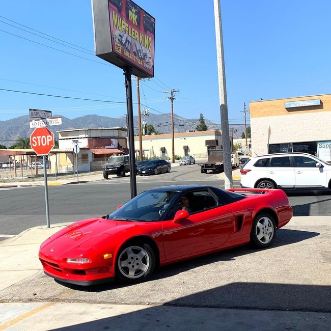 Red Honda NSX sports car parked on a street corner under a sign, with buildings and a mountain backdrop.
