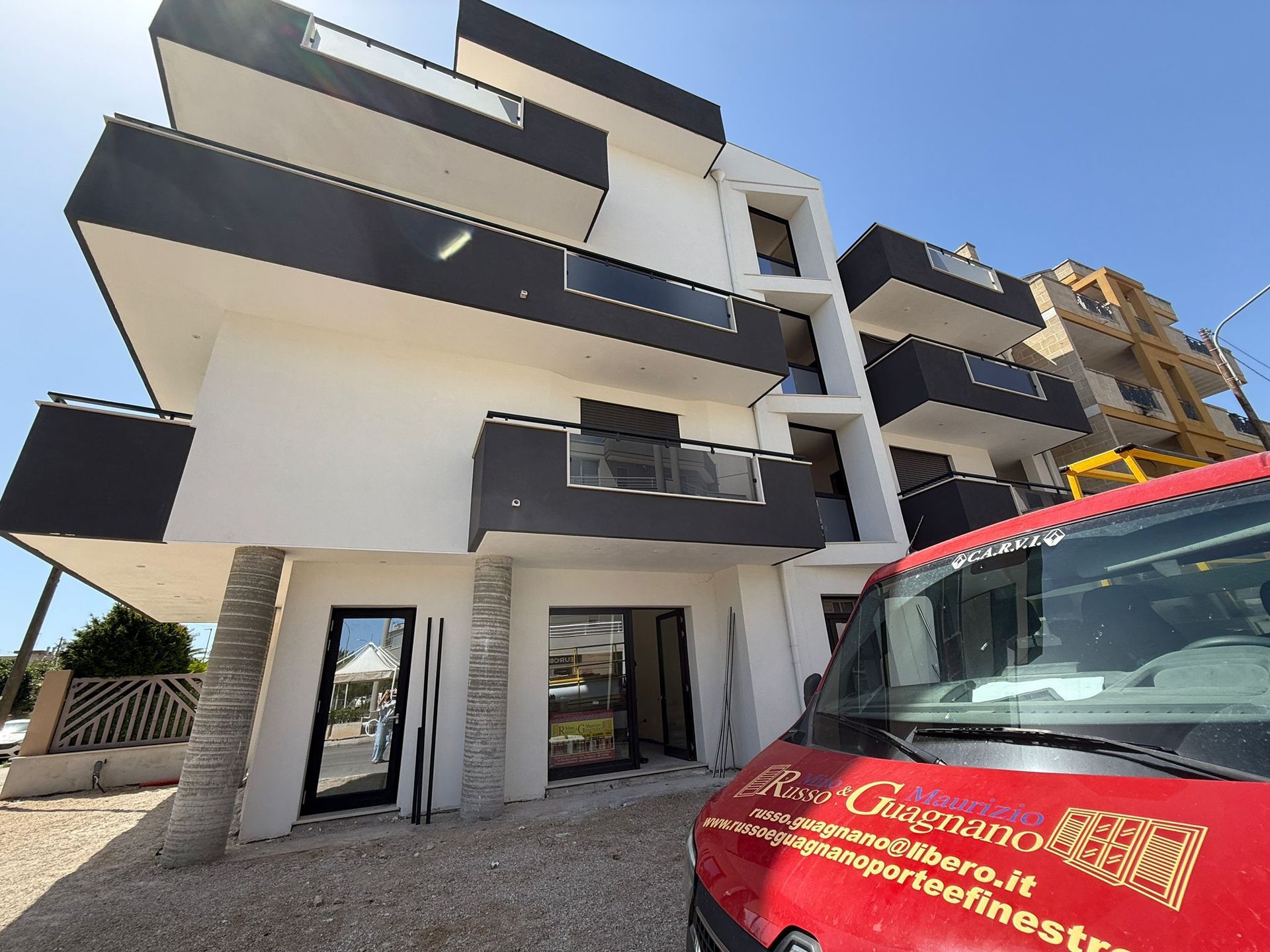 Moderno edificio bianco con balconi grigio scuro e furgone rosso davanti.