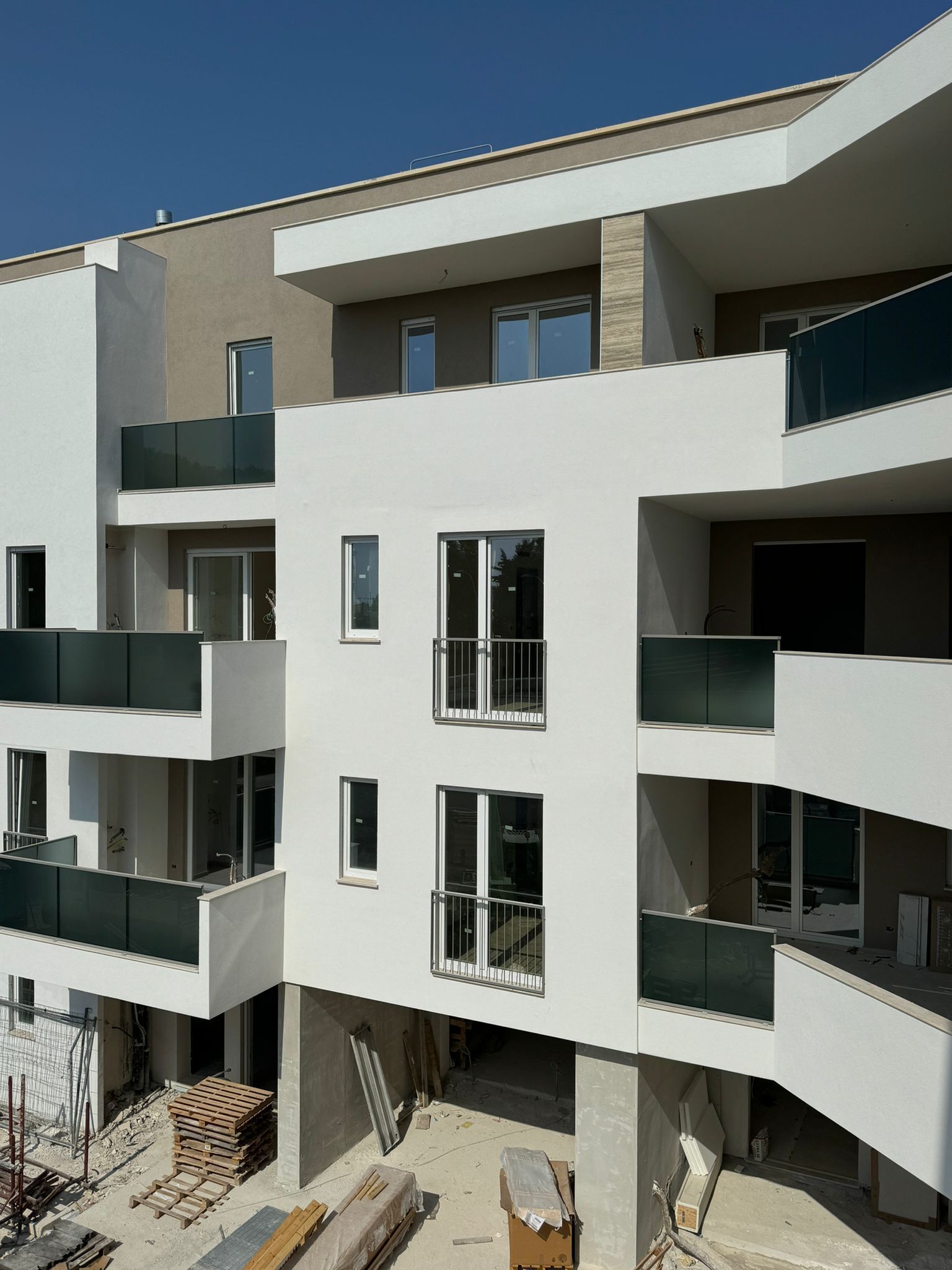 Moderno condominio in costruzione; esterni bianchi, balconi in vetro, travi in legno, cielo sereno.