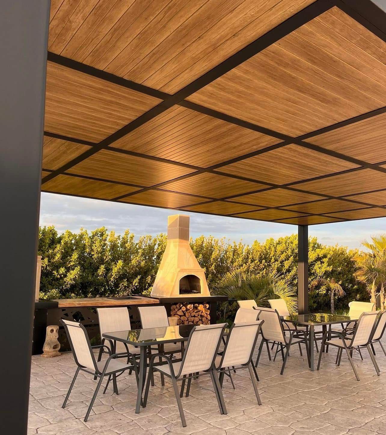 Zona pranzo all'aperto con tavoli, sedie e forno a legna sotto un pergolato in legno.
