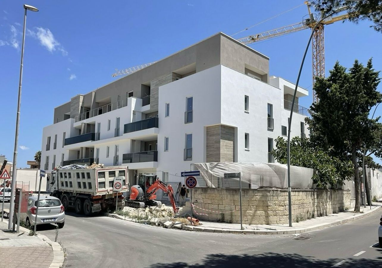 Cantiere con un edificio bianco e beige. Sono presenti un autocarro con cassone ribaltabile e un escavatore. Cielo azzurro.