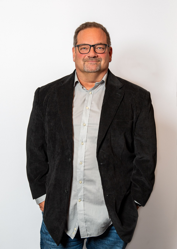 Standing in a black blazer and light shirt against a plain white background